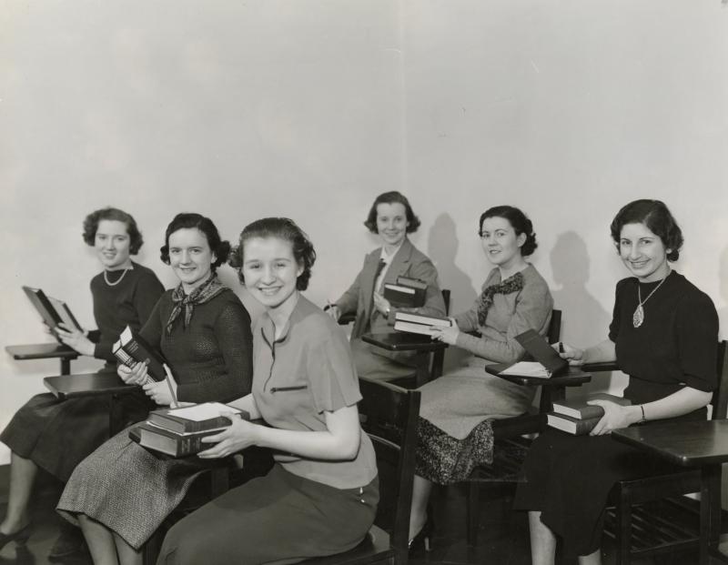 Six Mundelein College students sit at desks in a classroom, holding books and smiling at the camera.