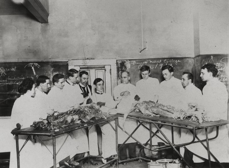 Ten students and a professor pose behind two operating tables in the Bennett Medical College Anatomy Lab. 