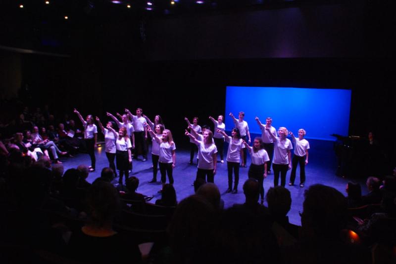 A group of Loyola University Chicago students in white t-shirts and black pants perform onstage, pointing out at the audience.