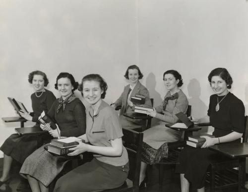 Six Mundelein College students sit at desks in a classroom, holding books and smiling at the camera.