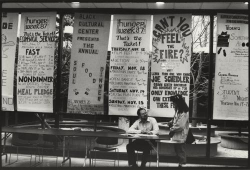 A man is sitting a table and talking to a standing women. There are six large "Hunger Week" posters behind them. 