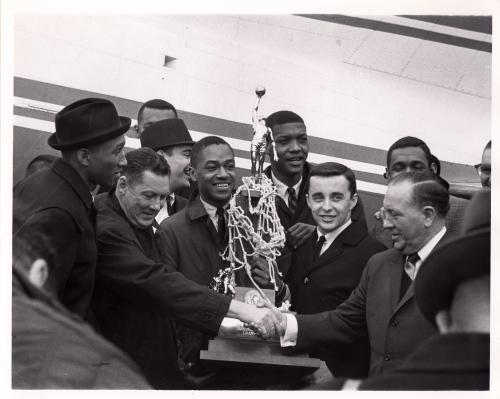 Mayor Richard Daley greets the 1963 team at the airport. The championship trophy, draped in a basketball net, is held between them. 