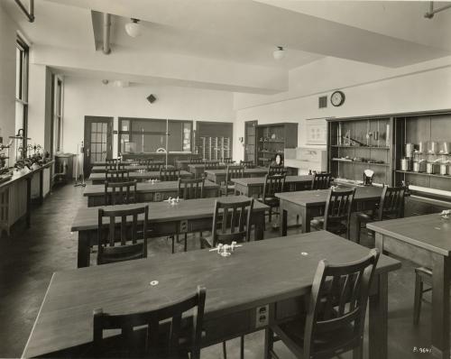 A shot of the chemistry lab at Mundelein College. There are rows of wooden tables and chairs in the center of the room, and shelves line the walls.